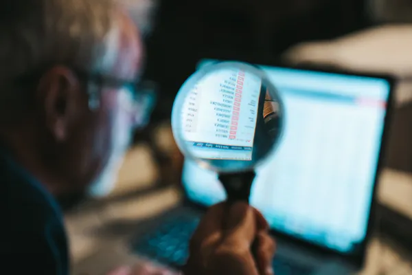 Un hombre sostiene una lupa frente a una pantalla con información financiera. Esta acción simboliza la búsqueda minuciosa de filtración de información.